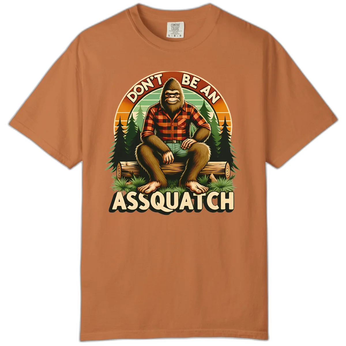 Vintage badge design with friendly Bigfoot in plaid shirt sitting on a log with forest trees, text reads 'Don't Be An Assquatch' in Yam